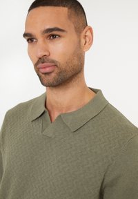Polo shirt i olivengrøn strik med et struktureret zigzag mønster, udstyret med en krave med v-halsudskæring og korte ærmer.
