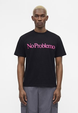 Mand med kort blondt hår iført sort t-shirt med pink "No Problemo" tekst og grå cargo bukser, står mod en hvid baggrund.