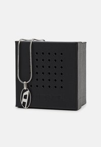 Silberne Anhänger-Halskette mit ovalem Design, das einen ausgeschnittenen "A" zeigt, an einer silbernen Kette, präsentiert neben einer schwarzen strukturierten Box.