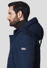 Granatowa kurtka zimowa z kapturem, wykonana z teksturowanej tkaniny. Posiada zamek błyskawiczny z przodu, boczne kieszenie oraz naszywkę z logo na lewym rękawie.
