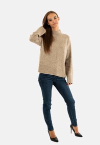 Suéter beige de punto con cuello alto, corte holgado y dobladillo acanalado, combinado con jeans skinny azul oscuro y tacones negros.