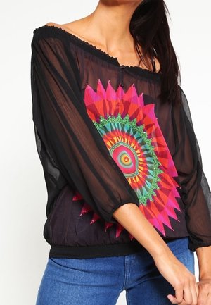 Vrouw draagt een doorschijnende zwarte off-shoulder blouse met een kleurrijk rond mandaladesign en blauwe spijkerbroek.