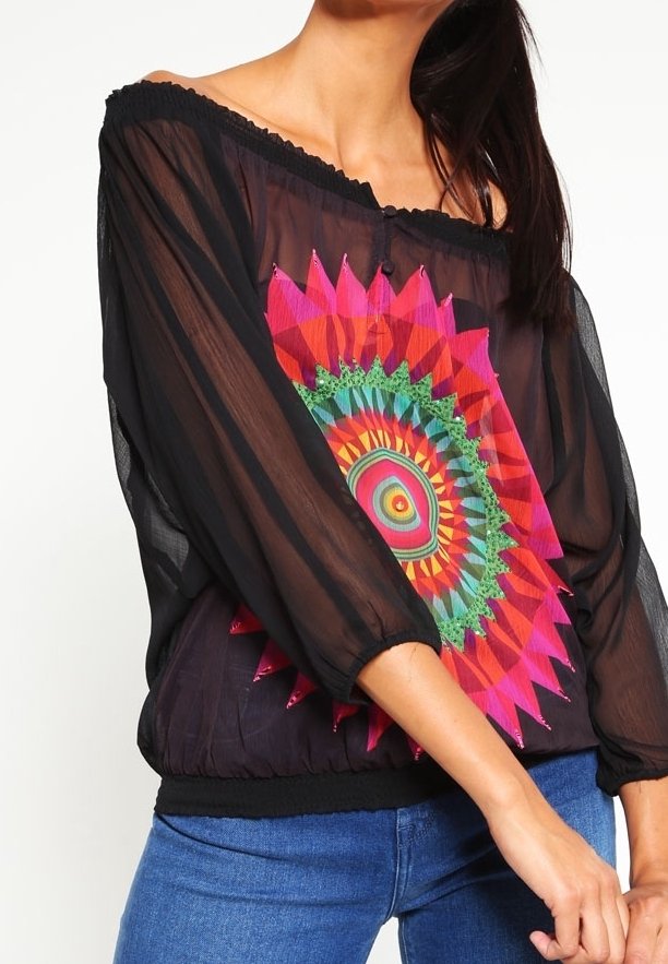 Femme portant un chemisier noir transparent à épaules dénudées avec un motif mandala circulaire coloré et un jean bleu.