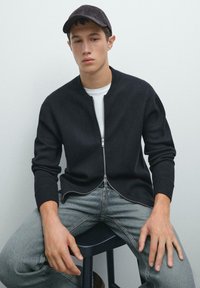 Svart zip-up cardigan med rund hals, ribbade ärmar, som bärs över en vit t-shirt, i kombination med ljusa denimjeans och en mörk keps.