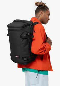 Mochila negra con un diseño rectangular, hecha de tejido duradero. Cuenta con correas ajustables, hebillas y un parche con el logo en la parte frontal.