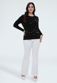 Frau in schwarzem Langarmshirt mit goldenen Ösendetails und weißen, weit geschnittenen Hosen steht mit einer Hand in der Hüfte vor einem schlichten Hintergrund.