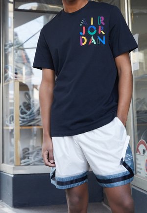 Czarna koszulka z kolorową grafiką "AIR JORDAN", zestawiona z białymi, siateczkowymi szortami z niebieskimi akcentami i paskowanymi ściągaczami.