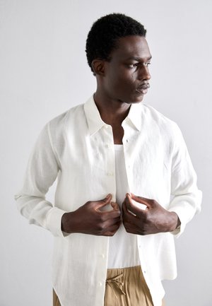 Weißes Leinenhemd mit Kragen, über einem hellen Tanktop getragen. Das Modell schließt die Knöpfe und trägt dazu beigen Hosen.