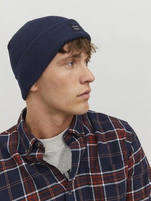 Navy ribbed knit beanie with a label, worn by a person in a plaid shirt featuring blue, red, and white colors. Close-up side view.