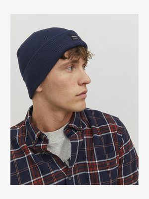 Marineblauwe geribde gebreide beanie met een label, gedragen door een persoon in een geruit overhemd met de kleuren blauw, rood en wit. Close-up zijaanzicht.