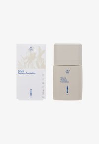 Base Natural Radiance en un envase minimalista beige con tapa estriada, acompañada de una caja blanca con un sutil diseño abstracto y texto azul.
