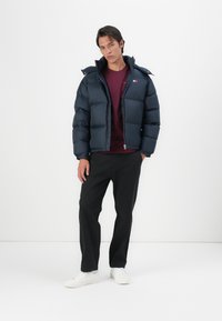 Mornarsko plavi puferjak s quilted dizajnom i kapuljačom, kombiniran s maroon košuljom i crnim hlačama. Bijele tenisice upotpunjuju odjevnu kombinaciju.