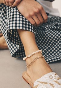 White beaded sandals with floral accents, paired with a delicate pink beaded anklet and a gold chain anklet, against a checkered fabric.