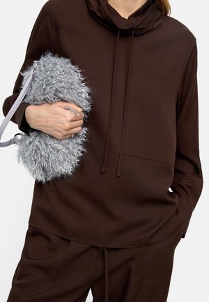 Osoba ubrana w ciemnobrązową bluzę z kapturem i spodnie, trzymająca małą, puszystą szarą torebkę z paskiem na nadgarstek na białym tle.
