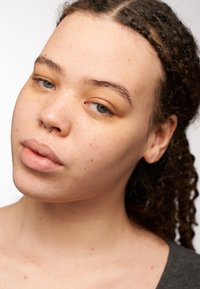 Gros plan sur une personne au teint lisse et brun clair, portant un piercing au septum en argent et ayant de longs cheveux bouclés et sombres.