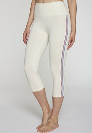 Woman wearing cream cropped leggings with red, white, and blue side stripes and "H.I.S" logo near left ankle, barefoot on light background.