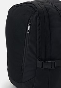 Mochila negra con un panel frontal texturizado, bolsillo lateral con cremallera, correa ajustable y cierre de hebilla para un almacenamiento seguro.