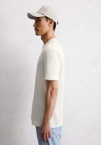 T-shirt en coton beige à manches courtes, encolure ronde et coutures d'épaule discrètes. Associé à un jean en denim bleu clair et une casquette beige.