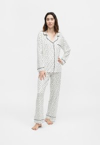 Νεαρή γυναίκα στέκεται ξυπόλητη, φορώντας λευκή πυτζάμα με κουμπιά και μακριά μανίκια, καθώς και παντελόνι με μαύρο τρίγωνο σχέδιο και ρέλι.