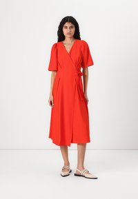 Rochie roșie cu mâneci scurte tip bulă, tiv până la genunchi și detaliu cu fundă pe lateral. Material neted cu o ușoară strălucire. Asortată cu balerini de culoare deschisă.