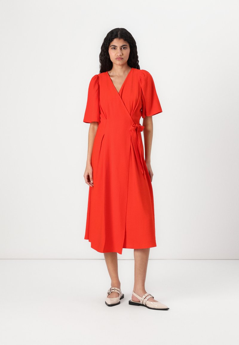Rochie roșie cu mâneci scurte tip bulă, tiv până la genunchi și detaliu cu fundă pe lateral. Material neted cu o ușoară strălucire. Asortată cu balerini de culoare deschisă.
