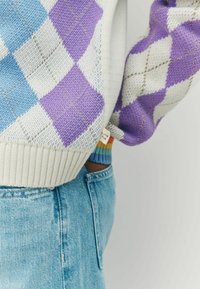 Sweter dziergany w diamentowy wzór w odcieniach niebieskiego i purpurowego, z prążkowanym, kremowym dołem i kolorowymi paskami na mankietach. W połączeniu z jasnymi dżinsami.