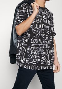 Černé oversized tričko s výraznými bílými textovými vzory, včetně "Versace Jeans Couture," v kombinaci s tmavými kalhotami a černou bundou.