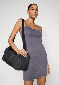 Juoda, tekstūruota kelioninė kuprinė su apvaliu pavidalu, reguliuojama petneša ir viršutiniu užtrauktuku. Moteriška modelis vilki prigludusią, pilkai-purpurinę liemenę.