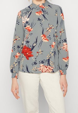 Blouse florale en tissu gris clair, ornée de fleurs rouges et pêches avec des feuilles vertes. Col haut avec détail froncé, manches longues avec poignets élastiques.