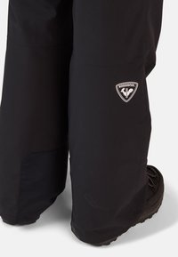 Zwarte waterdichte sneeuwbroek met een gladde textuur, voorzien van een Rossignol-logo op de dij en een versterkt ondergedeelte.
