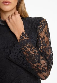 Black lace top featuring long, sheer lace sleeves with floral patterns and scalloped edges. Solid black bodice with a high neckline.