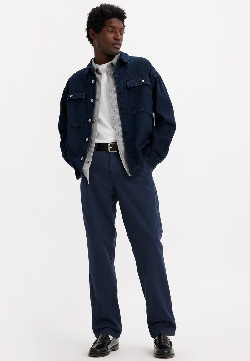 Chaqueta de mezclilla en azul oscuro, con dos bolsillos en el pecho y botones a presión. Debajo, una camisa gris claro, combinada con pantalones azul marino y zapatos negros.