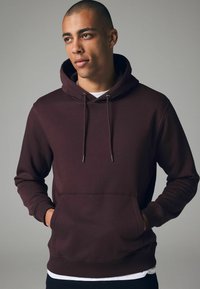 Sudadera burdeos hecha de tela suave, con un bolsillo frontal, cordones y puños de canalé. El modelo está de pie con las manos en los bolsillos.