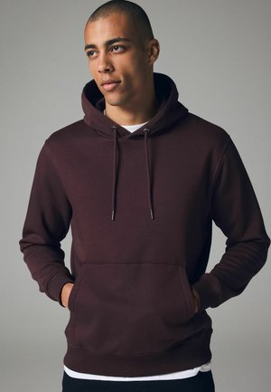 Maroon hoodie made from soft fabric, featuring a front pocket, drawstrings, and ribbed cuffs. Model stands with hands in pockets.