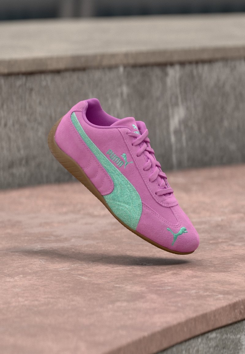 Baskets Puma en daim rose avec logo et bande bleu sarcelle, semelle marron, présentées en l’air au-dessus d’une surface en pierre et d’un fond en béton.