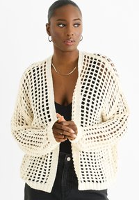 Open-knit cream cardigan made of textured yarn. Features a loose fit, long sleeves, and ribbed cuffs, worn over a black top.