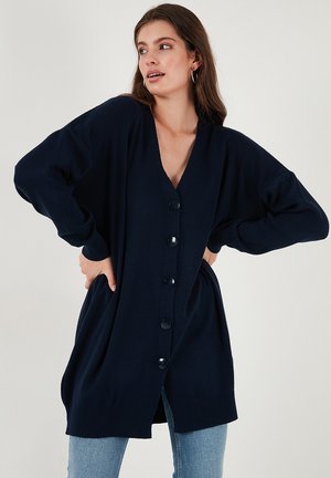 Donna in un cardigan oversized blu navy con bottoni e tasche, che indossa jeans azzurri, posando con le mani sui fianchi, guardando a destra.