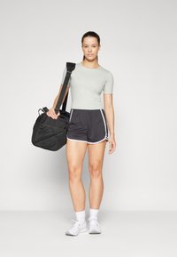 Lys grønn cropped t-skjorte, svarte treningsshorts med hvit kant, og hvite sneakers. Holder en svart duffel bag med skulderstropp.