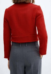 Red cropped jacket with textured fabric, frayed hem, and long sleeves, paired with gray trousers featuring a back pocket.