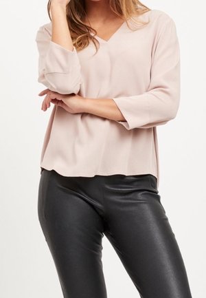 Woman wearing a light pink V-neck blouse with three-quarter sleeves and black faux leather pants, arms crossed at the waist.