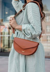 Hnědá kožená crossbody taška s zakřiveným klopovým uzávěrem a zlatými detaily, která má vyražené logo. Popruh je nastavitelný.
