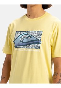 Camiseta de algodón amarilla con un logotipo gráfico azul y un patrón texturizado en el frente. Diseño de manga corta y cuello redondo.