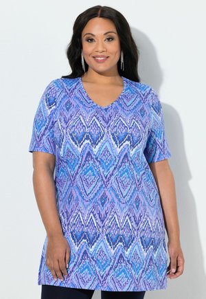 Kobieta ubrana w tunikę w niebiesko-fioletowy wzór diamentowy z dekoltem w serek i krótkimi rękawami, stojąca na białym tle.