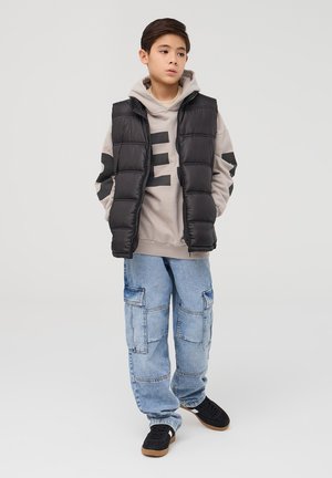 Gilet nero a soffietto su felpa beige con grande scritta nera, abbinato a jeans cargo blu chiaro e sneakers nere. Sfondo semplice.