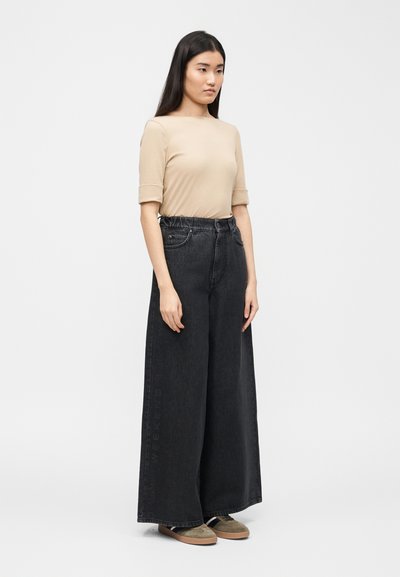 Γυναίκα με μακριά σκούρα μαλλιά που φοράει μπεζ κοντομάνικη μπλούζα και μαύρο τζιν παντελόνι με ψηλή μέση και φαρδιά μπατζάκια, στέκεται μπροστά σε απλό ανοιχτό φόντο.