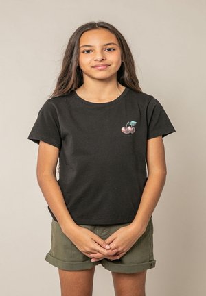 Fille aux longs cheveux bruns portant un t-shirt noir avec un motif de cerise et un short vert retroussé, debout les mains jointes.