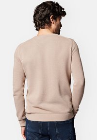 Beżowy sweter dzianinowy o teksturowanym wzorze, z prążkowanymi rękawami i dołem oraz klasycznym okrągłym dekoltem. Noszony z ciemnoniebieskimi dżinsami.