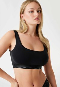 Bralet negru cu un design ajustat, bretele late și bandă elastică cu logo-ul „GUESS”. Textură netedă și formă conturată.