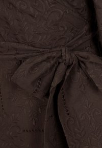 Brązowy teksturowany materiał z misternym haftem kwiatowym, związany w talii kokardą, podkreślający szczegółowy wzór odzieży.