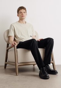 Ljusbeige t-shirt med en svart logotyp, svarta byxor och blanka svarta skor, sittande på en stol klädd i ljus tyg med trästomme.